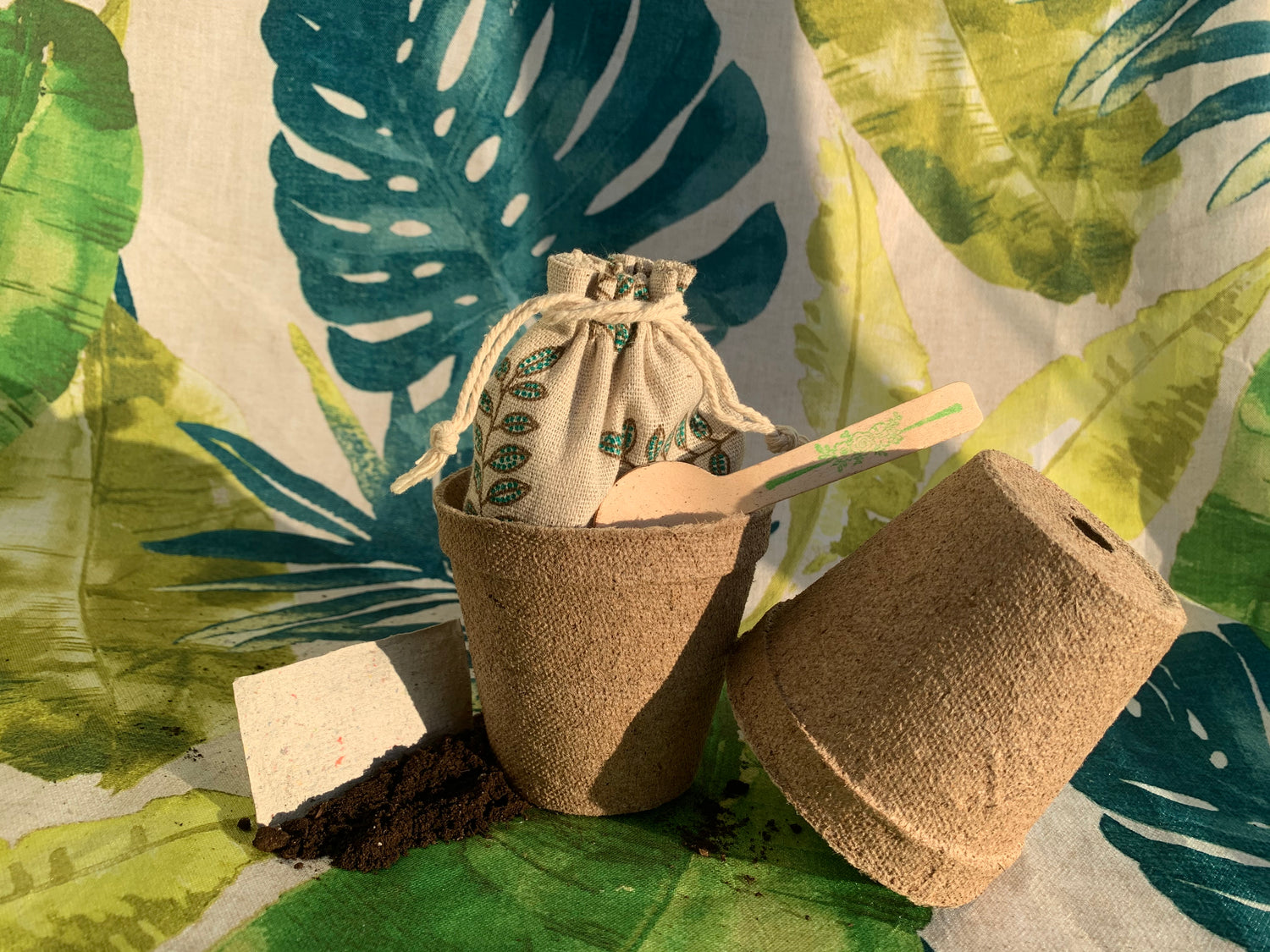 Soil with seedpaper ontop. Two pots, one with a pounch and spoon inside. Background with leafs.